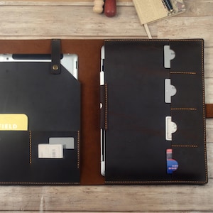 May include: A brown leather portfolio with a black leather interior. The portfolio has a pocket for a tablet, a pocket for a notepad, and several card slots. The portfolio is closed and lying flat on a wooden surface.