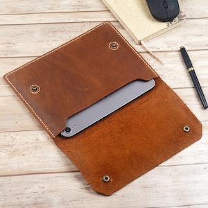 Peut inclure: Une pochette pour tablette en cuir marron avec une fermeture à rabat et des accents métalliques. La pochette est ouverte et une tablette est visible à l'intérieur.
