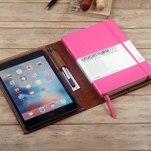 May include: A brown leather folio case with a pink notebook and a tablet computer inside. The notebook has the text "LEUCHTTURM1917 DETAILS THAT MAKE ALL THE DIFFERENCE" on the cover.