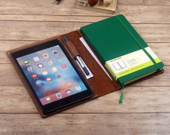 Könnte beinhalten: Ein braunes Lederetui mit einem grünen Moleskine Classic Notizbuch und einem Tablet-Computer. Das Etui hat einen Stifthalter und einen Platz für ein Tablet.
