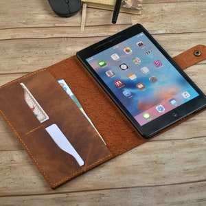 Op de afbeelding: Een bruine leren tablet-hoes met een flap die over de tablet vouwt om deze te beschermen. De hoes heeft een gleuf voor kaarten en een vak voor contant geld. De tablet is zichtbaar in de hoes en heeft een scherm met verschillende apps die worden weergegeven.