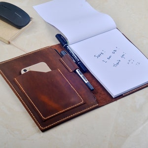 Peut inclure: Une couverture de carnet en cuir marron avec un porte-stylo et une poche pour un téléphone. Le carnet est ouvert et a une page blanche avec des notes manuscrites qui disent "Bien sûr ! Je vais bien ! Merci !"