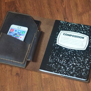 May include: A brown leather notebook cover with a pocket for a phone and a pen holder. The cover is open to reveal a black composition notebook with the word "COMPOSITION" printed on the front.