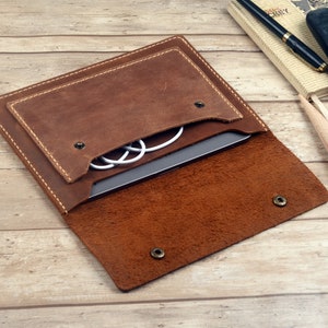 Pode incluir: Uma capa para laptop de couro marrom com uma aba que se dobra e fecha com um botão de pressão. A capa tem um bolso na frente para guardar cabos ou outros itens pequenos. A capa está aberta e um laptop e cabos são visíveis no interior.
