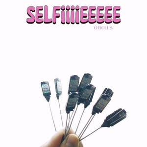 Puede incluir: Una mano sostiene un ramo de teléfonos móviles en miniatura sobre palos. Los teléfonos son negros y tienen una pantalla que muestra una imagen genérica. El texto "SELFiieeeeee" y "ó DOLLS" está escrito en rosa sobre los teléfonos.