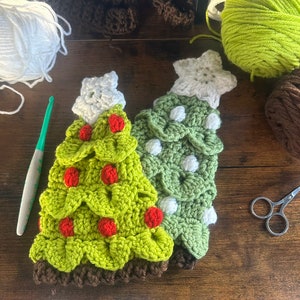 May include: Two crocheted Christmas tree ornaments, one green with white polka dots and a white star, and one green with red pom-poms. Both ornaments are on a wooden surface.