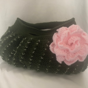Puede incluir: Una bolsa de crochet verde con una flor de crochet rosa unida al lado. La bolsa tiene una superficie texturizada y está hecha con un hilo grueso.