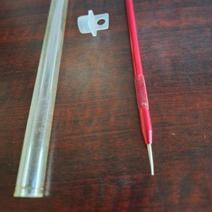 May include: A clear glass tube with a white plastic cap and a red metal tool with a pointed tip. The tool is used for crafting and art projects.