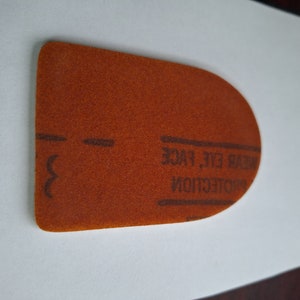 May include: A rust-colored, D-shaped sanding pad with the text "WEAR EYE, FACE PROTECTION" printed on it. The pad has a textured surface and a curved top edge. The number "3" is printed on the left side.