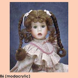 May include: A brown doll with long, braided hair and a white dress with pink accents. The doll has blue eyes and a sweet expression. The doll's hair is styled in two braids with white bows.