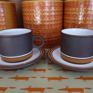 May include: Two brown coffee cups with matching saucers. Each cup has a white stripe and a tan band near the base. The cups and saucers are on a tray with a fox pattern. Orange biscuit jars are in the background.