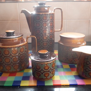 May include: A ceramic tea set featuring a tall coffee pot, a teapot, a lidded sugar bowl, a lidded tea canister with the word TEA embossed on the side, and a creamer. Each piece has a brown and teal circular pattern with a glossy finish.
