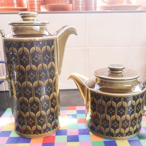 May include: Two ceramic teapots with a brown and gold geometric pattern. One is tall with a handle, the other is shorter with a lid. The teapots have a retro design and are likely vintage.