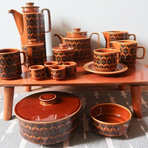 May include: A collection of brown ceramic tableware featuring a repeating dark geometric pattern. The set includes a tall coffee pot, a teapot, a lidded sugar bowl, a creamer, several mugs with saucers, small cups, and a large lidded serving dish.