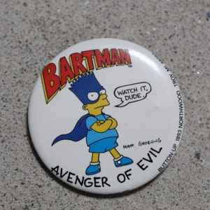 May include: A white button with a cartoon image of Bart Simpson dressed as a superhero. The button says "Bartman" in red and yellow letters, "Avenger of Evil" in black letters, and "Watch it, dude." in a speech bubble. The button also has the text "Button-Up 1893 Northwood Troy" and "Matt Groening" in smaller letters.