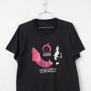 May include: Black t-shirt with a pink and white graphic of a musical note and the text "Sanford Symphony Musical Artistry The 1982-1983 Season"