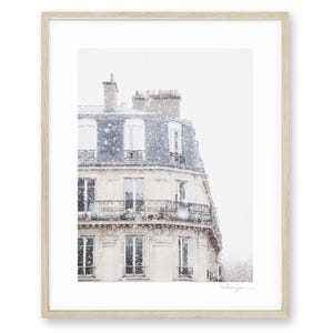 May include: A framed print depicting a Parisian building in winter. The building features a light-coloured facade, a dark blue roof, and wrought iron balconies. Gentle snowfall creates a serene, wintry scene. The frame is light wood.