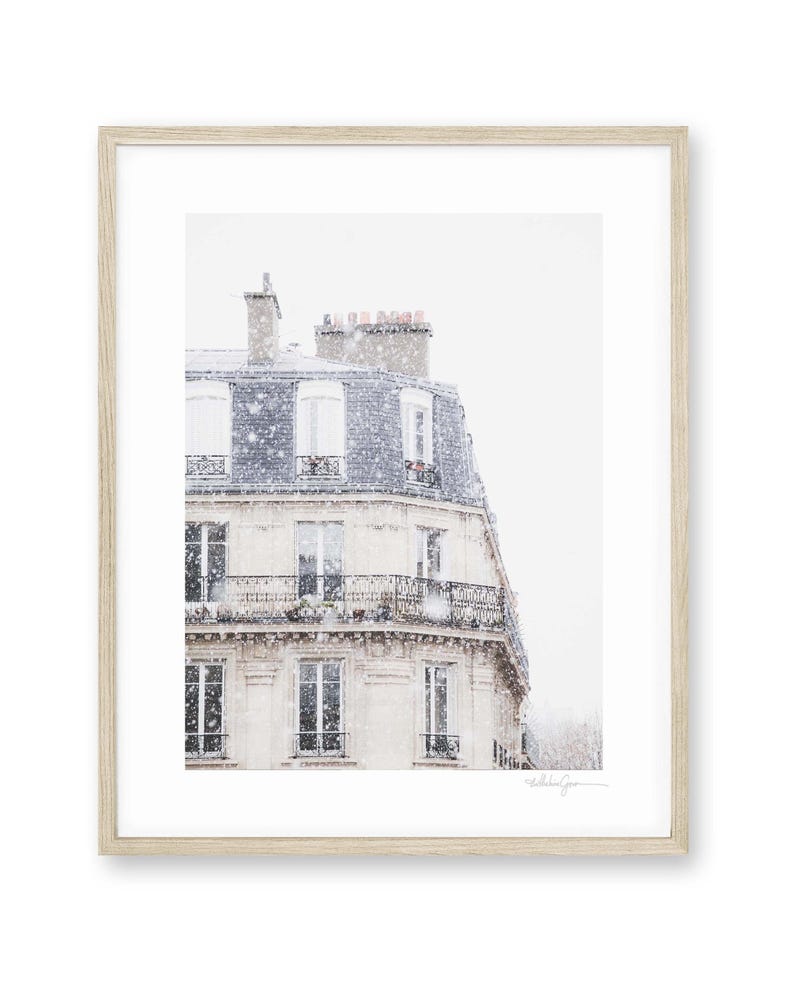 May include: A framed print depicting a Parisian building in winter. The building features a light-coloured facade, a dark blue roof, and wrought iron balconies. Gentle snowfall creates a serene, wintry scene. The frame is light wood.