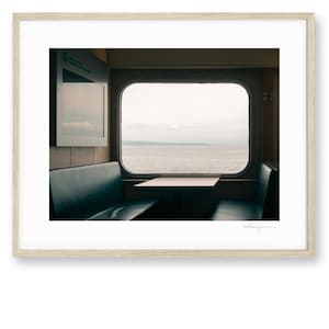 Op de afbeelding: Een ingelijste foto met uitzicht vanuit een veerbootraam over kalm water onder een bewolkte lucht. Het interieur toont donkerblauwe banken aan weerszijden van een kleine tafel, gevat in een lichtgekleurde houten lijst.