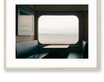 May include: A framed photograph depicting a view from a ferry window looking out over a calm body of water under a cloudy sky. The interior features dark blue bench seating on either side of a small table, with a light-colored wooden frame.
