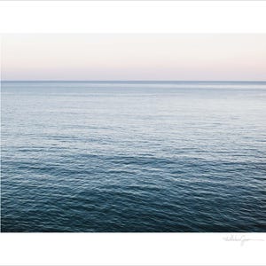 May include: A serene seascape print featuring a gradient of colors. The image showcases the ocean, transitioning from deep blue at the bottom to lighter shades of blue and a soft pinkish-white sky at the top. The water's surface has gentle ripples.