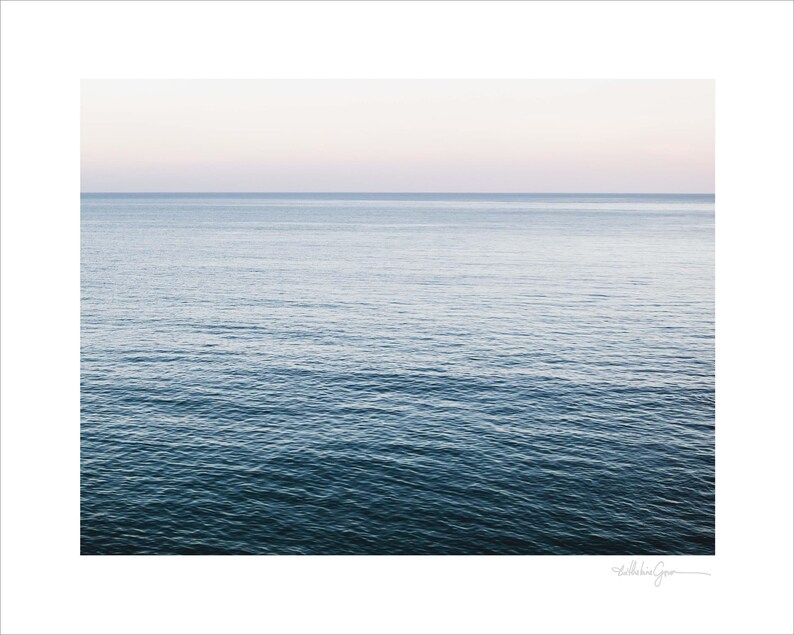 May include: A serene seascape print featuring a gradient of colors. The image showcases the ocean, transitioning from deep blue at the bottom to lighter shades of blue and a soft pinkish-white sky at the top. The water's surface has gentle ripples.