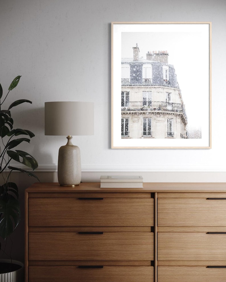 May include: A framed print of a Parisian building in a snowstorm. The print is displayed above a wooden chest of drawers with a lamp and a plant. The lamp has a beige shade and a ceramic base.