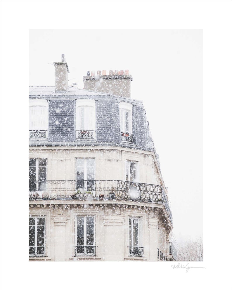 May include: A photograph of a Parisian building in a snowstorm. The building has a light-coloured facade with multiple windows and balconies. The roof is a dark grey colour. Snowflakes are falling, creating a winter scene.