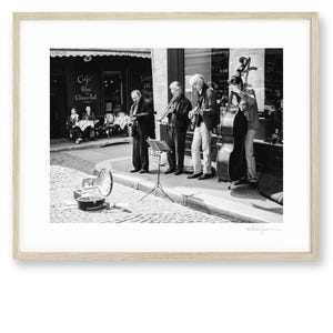 Op de afbeelding: Zwart-wit foto in een lijst van een straatbeeld met een jazzband die instrumenten bespeelt. De band bestaat uit een saxofoon, gitaar, viool en contrabas. Op de achtergrond is een café met de tekst "Cafe du Chocolat" te zien.