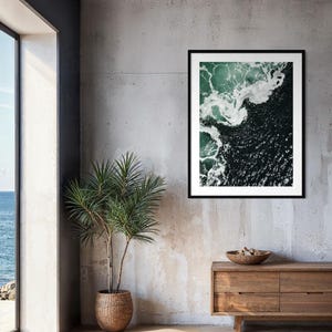 Op de afbeelding: Een ingelijste kunstdruk met een luchtfoto van oceaangolven. Het kunstwerk toont donkergroene en witte waterpatronen, in een zwart frame. De print hangt aan een betonnen muur boven een houten kast. Een potplant is ook zichtbaar.