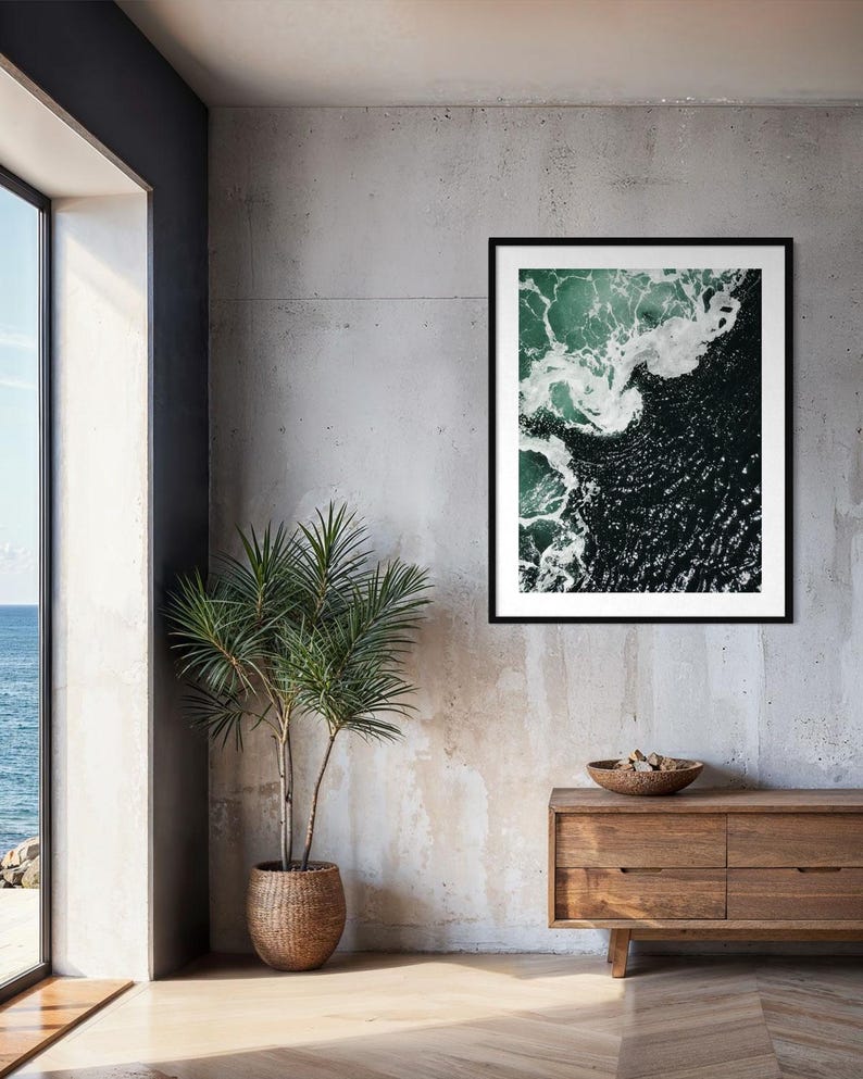 Op de afbeelding: Een ingelijste kunstdruk met een luchtfoto van oceaangolven. Het kunstwerk toont donkergroene en witte waterpatronen, in een zwart frame. De print hangt aan een betonnen muur boven een houten kast. Een potplant is ook zichtbaar.