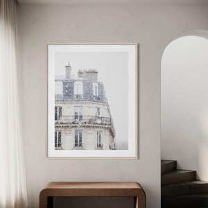 May include: A framed print depicting a Parisian building during a snowfall. The building features multiple floors with balconies and windows. The print is in a light wood frame, set in a neutral room with a brown bench.