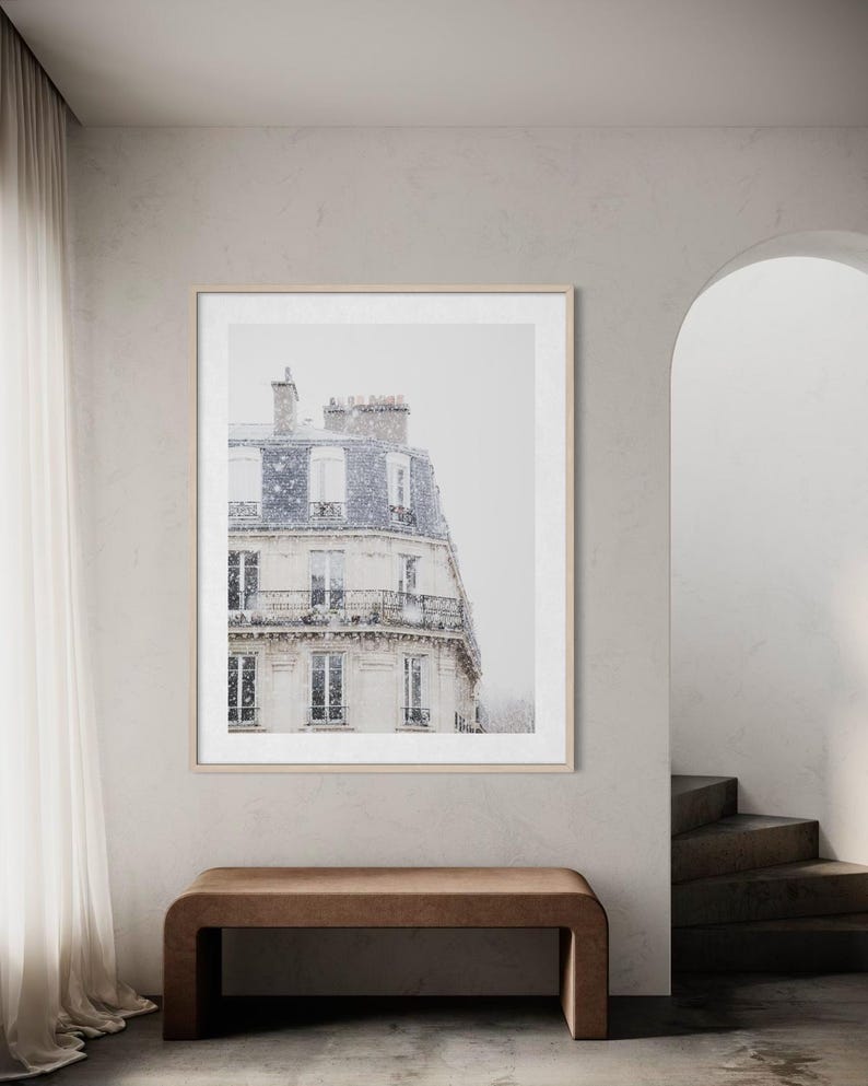 May include: A framed print depicting a Parisian building during a snowfall. The building features multiple floors with balconies and windows. The print is in a light wood frame, set in a neutral room with a brown bench.