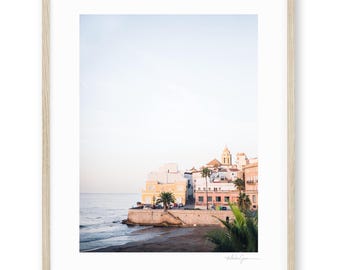 May include: A framed print featuring a coastal town scene. The image showcases buildings in shades of yellow, white, and pink along the shoreline. Palm trees and a church tower are visible. The sky is a soft, pale blue.