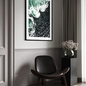 Op de afbeelding: Een ingelijste kunstdruk met een luchtfoto van oceaangolven. Het kunstwerk toont donkergroen en wit water, omlijst door een zwarte rand. Een zwarte leren stoel en een marmeren sokkel met een vaas bloemen staan op de voorgrond.