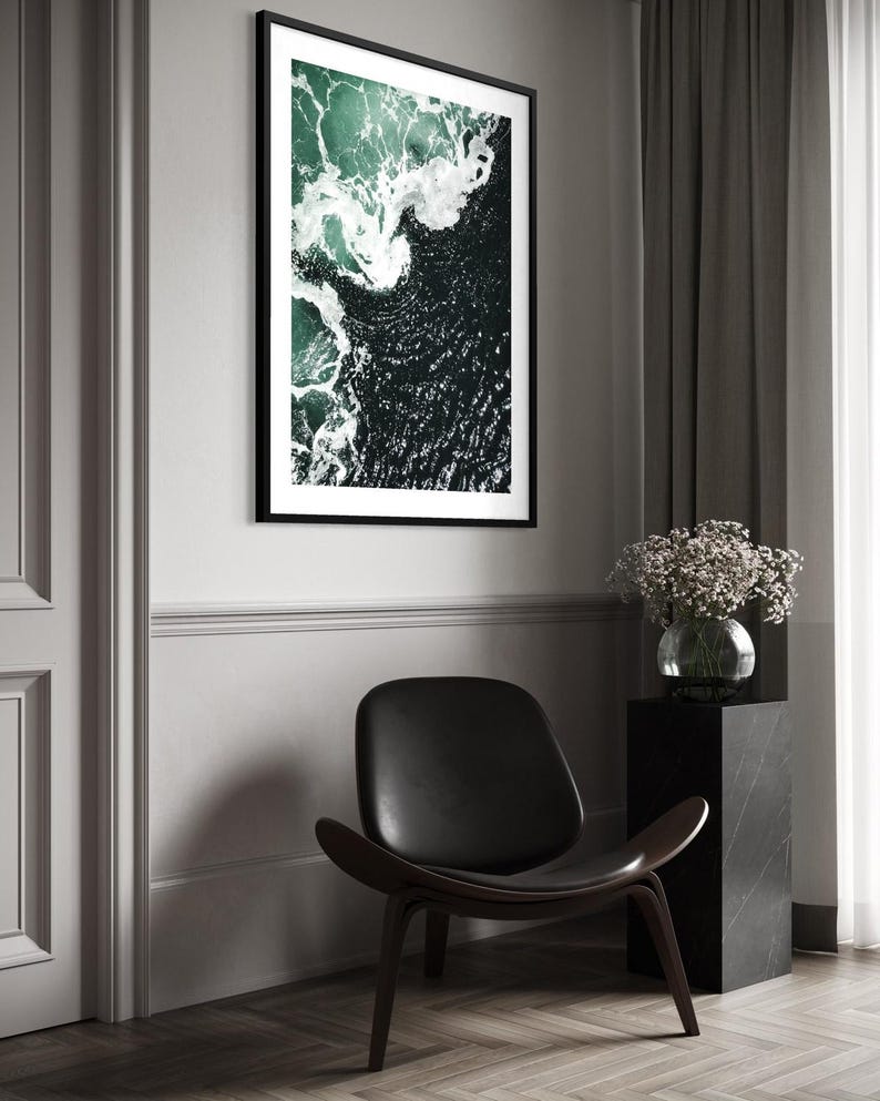 Op de afbeelding: Een ingelijste kunstdruk met een luchtfoto van oceaangolven. Het kunstwerk toont donkergroen en wit water, omlijst door een zwarte rand. Een zwarte leren stoel en een marmeren sokkel met een vaas bloemen staan op de voorgrond.