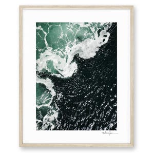 Op de afbeelding: Abstracte ingelijste print met een luchtfoto van oceaangolven. De afbeelding toont donkerblauwgroen water met witte schuimende golven. Het kunstwerk is ingelijst in een licht houten frame. De print is rechtsonder gesigneerd.