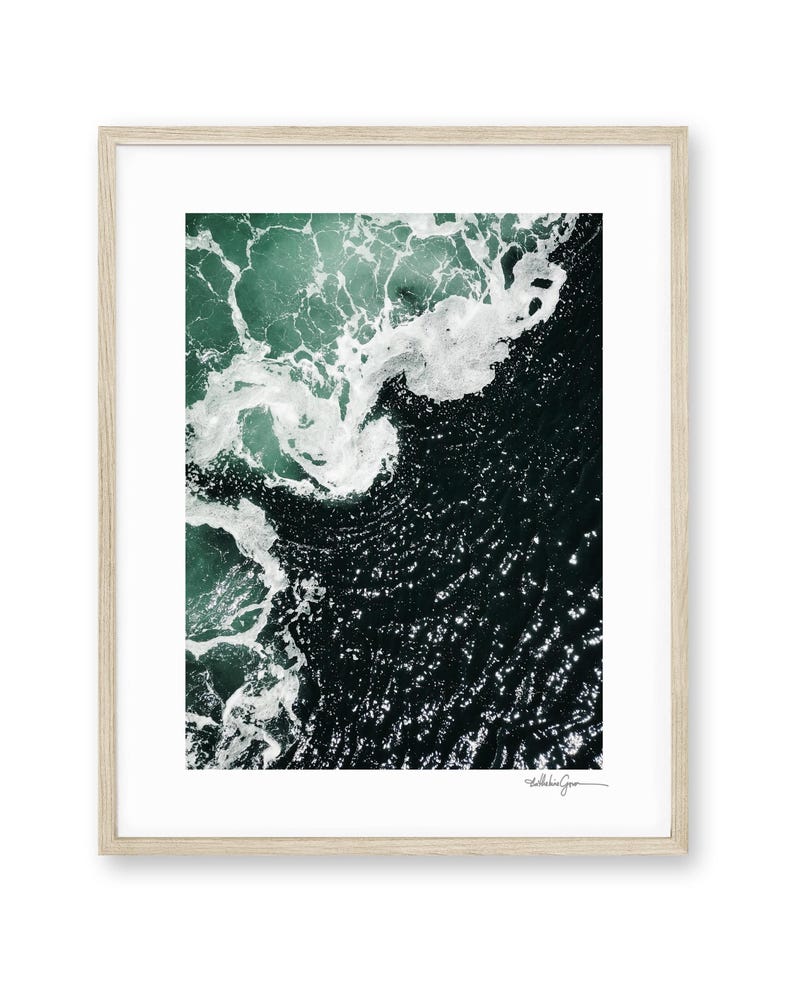 Op de afbeelding: Abstracte ingelijste print met een luchtfoto van oceaangolven. De afbeelding toont donkerblauwgroen water met witte schuimende golven. Het kunstwerk is ingelijst in een licht houten frame. De print is rechtsonder gesigneerd.