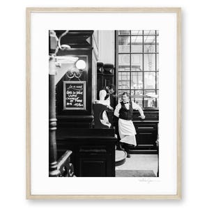 Op de afbeelding: Een zwart-wit ingelijste foto van twee obers in een klassieke bistro. De ene ober staat bij een krijtbord met handgeschreven tekst, terwijl de andere tegen een donkere houten toonbank leunt, gekleed in een gilet en een schort.