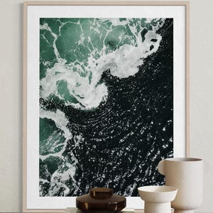Op de afbeelding: Een luchtfoto van oceaangolven in tinten teal en donkerblauw, met witte schuimende toppen. De afbeelding is omlijst in een licht houten frame. Decoratieve vazen en boeken staan op de voorgrond.