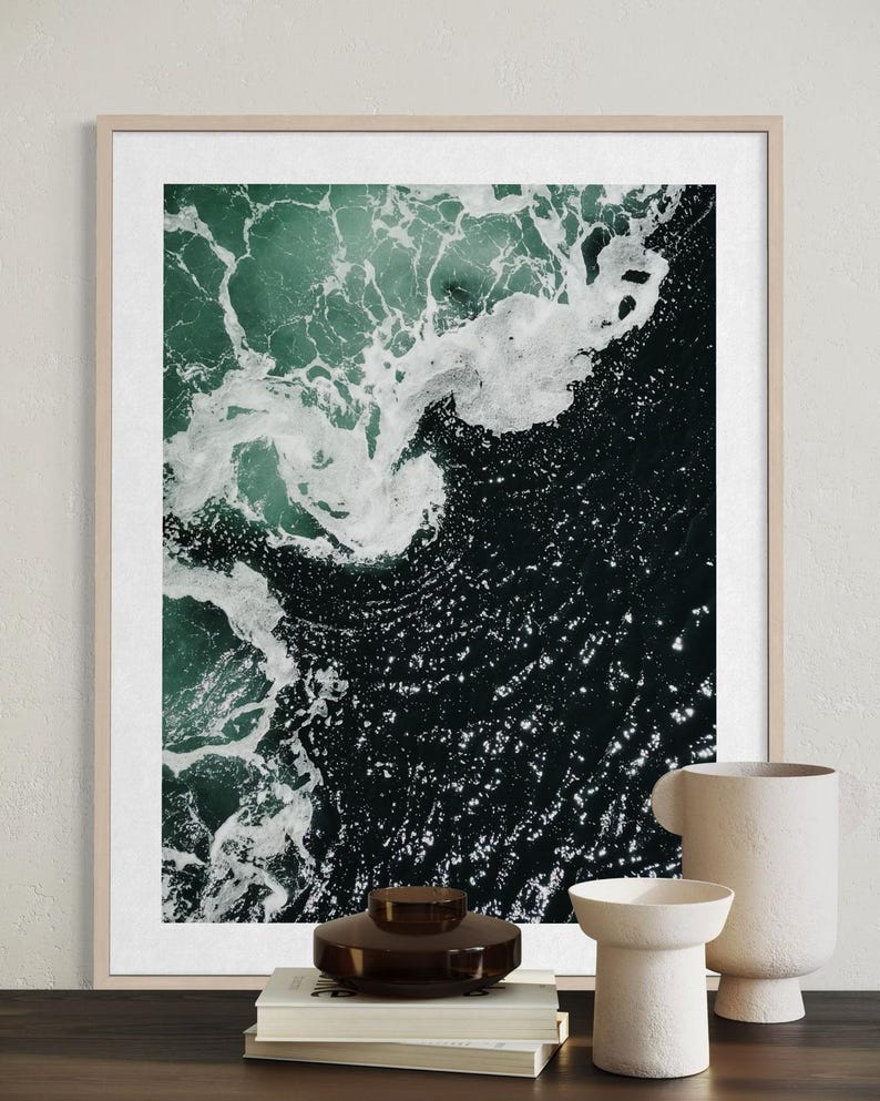 Op de afbeelding: Een luchtfoto van oceaangolven in tinten teal en donkerblauw, met witte schuimende toppen. De afbeelding is omlijst in een licht houten frame. Decoratieve vazen en boeken staan op de voorgrond.