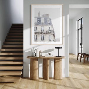 May include: A framed print of a Parisian building with a snow-covered roof hangs above a wooden console table. The table has three thick, cylindrical legs. A black lamp, books, and decorative vases sit on the table. A wooden staircase and hardwood floors complete the interior scene.