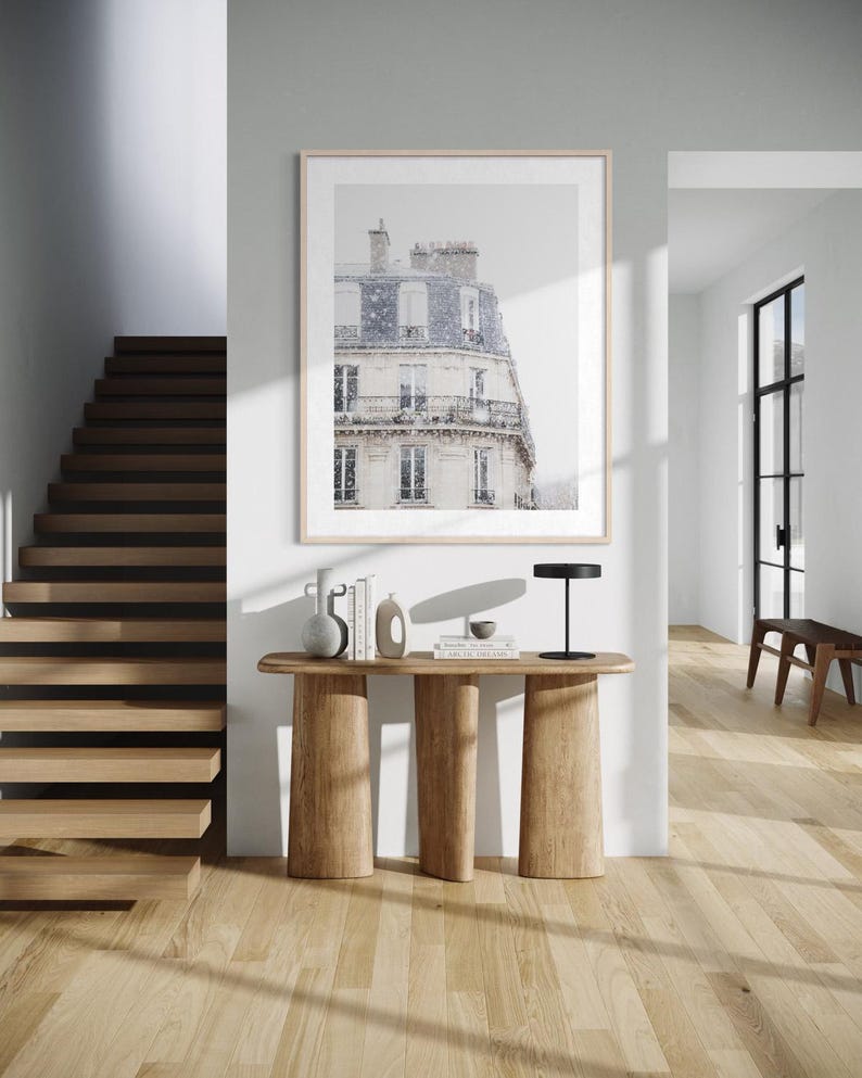 May include: A framed print of a Parisian building with a snow-covered roof hangs above a wooden console table. The table has three thick, cylindrical legs. A black lamp, books, and decorative vases sit on the table. A wooden staircase and hardwood floors complete the interior scene.