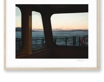 May include: A framed photograph captures a ferry's interior view, showcasing the ocean and mountains through a window. The scene is bathed in the warm hues of a sunset, with a life preserver visible. The image is framed in a light wood.