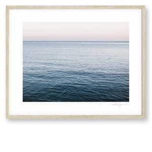 May include: A framed print featuring a serene seascape. The image captures the ocean's surface, transitioning from deep blue to a lighter hue near the horizon. The sky above fades from a soft pink to a pale blue. The frame is a light wood.
