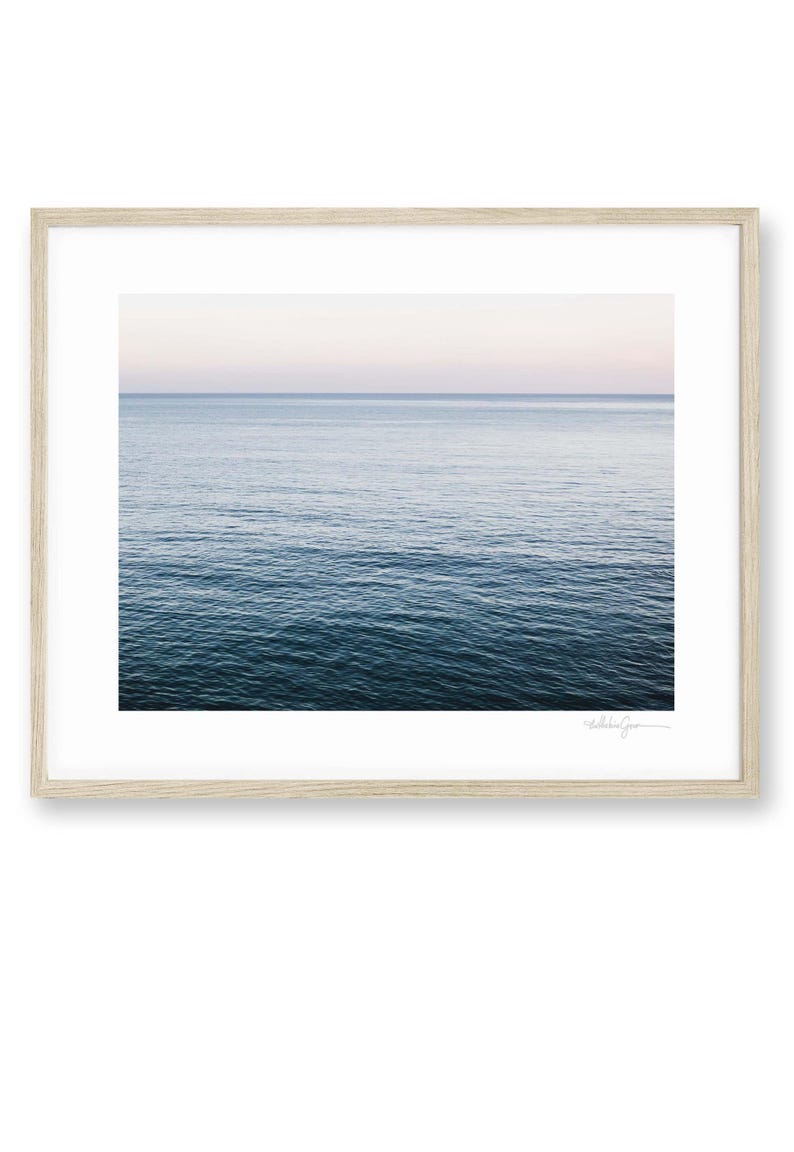 May include: A framed print featuring a serene seascape. The image captures the ocean's surface, transitioning from deep blue to a lighter hue near the horizon. The sky above fades from a soft pink to a pale blue. The frame is a light wood.