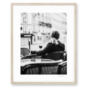 Op de afbeelding: Een zwart-wit ingelijste foto van een persoon van achteren gezien, zittend aan een ronde bistrotafel met een glas wijn. De scène is buiten met wazige stadsgebouwen op de achtergrond, geplaatst in een lichthouten lijst.