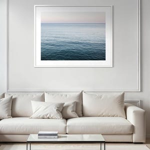 May include: A framed photograph of the ocean. The water transitions from a deep blue at the bottom to a lighter blue and pink at the horizon. The photograph is in a white frame and hangs above a white sofa and glass coffee table.