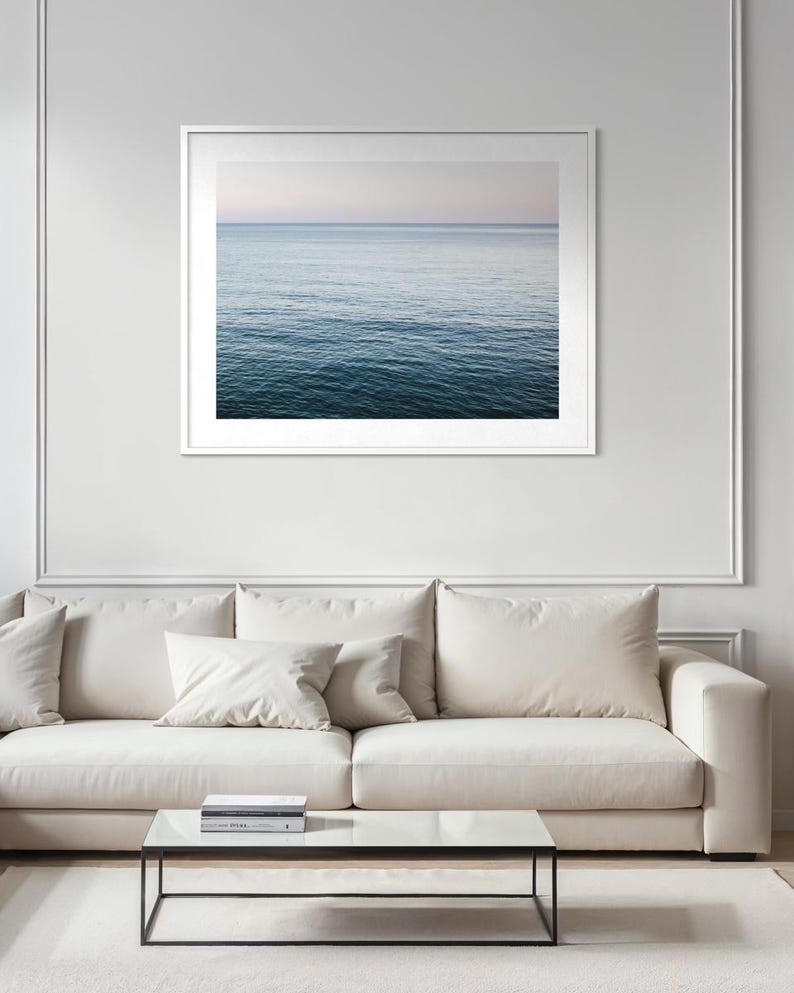 May include: A framed photograph of the ocean. The water transitions from a deep blue at the bottom to a lighter blue and pink at the horizon. The photograph is in a white frame and hangs above a white sofa and glass coffee table.