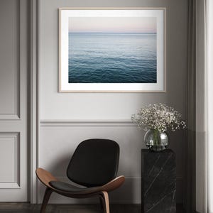 May include: A framed print of the ocean with a light wood frame. The water is a deep blue, and the sky is a soft pink and blue. A black chair with a wooden frame and a black marble pedestal with a glass vase of flowers are also in the image.